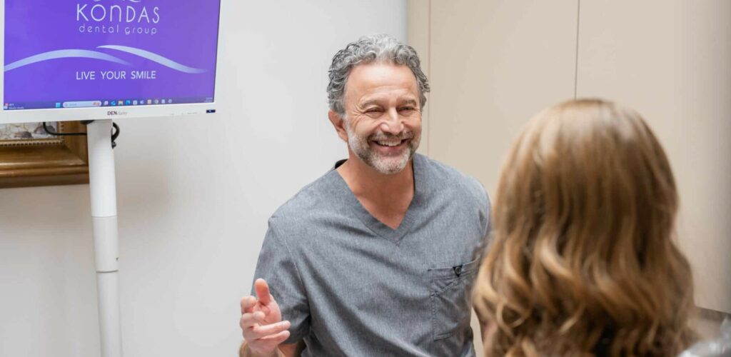 Smiling dentist in scrubs talks with a patient during an in-office dental consultation, with a wall-mounted monitor in the exam room.