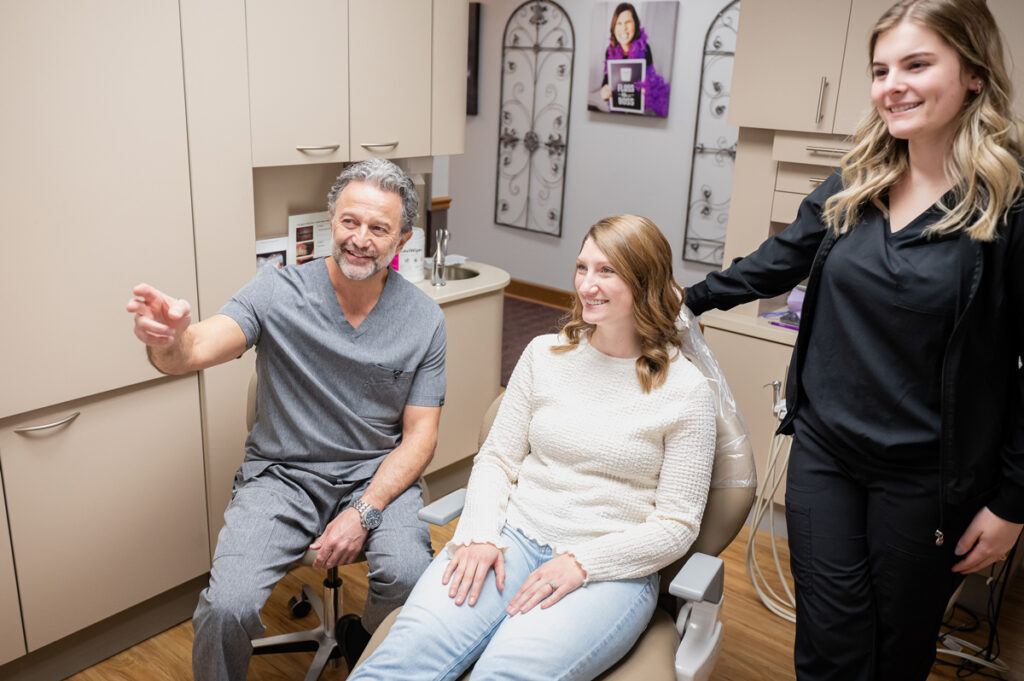 Dentist talks with a smiling patient in a dental chair while a dental assistant stands nearby in the exam room during a consultation.